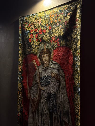 This is a close-up, eye-level, indoor shot of a tapestry hanging on a dark wall. The tapestry depicts an angel playing a harp. The angel has red wings and is wearing a grey robe. The background of the tapestry is a dense floral pattern with red, yellow, and blue flowers and green leaves. A spotlight illuminates the top portion of the tapestry, casting shadows on the upper right side. The tapestry is positioned in a dimly lit environment. There are no visible signs of the city of Бородино, Russia in the image.