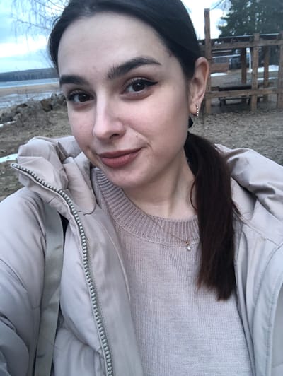 A woman is posing for a selfie in what appears to be an outdoor setting, possibly near the city of Kizhi, Russia, although specific Kizhi architectural elements aren't clearly visible. The woman is wearing a light pink sweater, a beige puffer jacket, and a delicate necklace with a small pendant. She has dark hair pulled back in a ponytail and is wearing eyeliner. In the blurred background, a wooden structure can be seen, potentially a fence or building. The ground appears to be covered with gravel and possibly some snow. In the distance, what looks like water or a snowy field is visible.