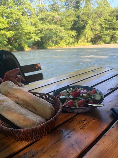 In Abzhanda, Abkhazia, a close-up shot captures a rustic wooden table set with a meal beside a flowing river. In the foreground, a woven basket holds two elongated loaves of bread. To the right, a bowl contains a vibrant salad composed of chopped tomatoes, cucumbers, and herbs, with a golden fork resting on its rim. A person's hand, holding a coral-colored smartphone, is visible on the left side of the frame, partially obscured by the basket. In the background, the wide, rippling river flows serenely, bordered by lush green trees and foliage under a bright sky.