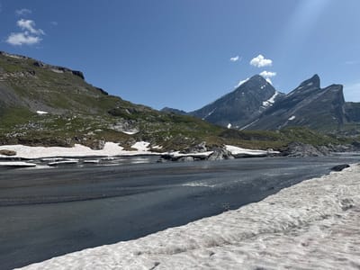 This is a landscape photograph taken in Leukerbad, Switzerland, depicting a mountainous scene with a river and snow. The sky is clear and blue, with a few wispy clouds. In the foreground, a wide, shallow river flows from left to right across the frame. The riverbed is composed of dark pebbles and sand. Snow is visible along the right bank of the river and in patches on the left bank, interspersed with rocky terrain and sparse green vegetation.
The middle ground features a rocky, green-covered mountainside rising steeply on the left, leading to a rugged ridge line. On the right, majestic, sharp-peaked mountains dominate the skyline. These peaks are dark gray rock with streaks of snow clinging to their upper slopes and crevices.
The overall environment suggests a high-altitude location during a season where snow is melting and rivers are active, likely late spring or early summer in the Swiss Alps. There are no people or animals visible in the image. The scene is natural and undeveloped.