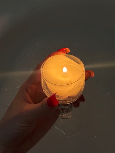 A human hand, likely female, with long red-painted fingernails, holds a clear glass stemmed goblet containing a lit white candle. The candle's flame is bright, casting a warm yellow-orange light that illuminates the hand, the wax, and the inner surface of the goblet, which shows solidified, irregular wax patterns. The goblet has a faceted base and stem. The background is dark and indistinct, revealing faint, blurred highlights consistent with the surface of water with small bubbles or foam, suggesting a bath environment. The scene is located in Чистенское сельское поселение, Ukraine.