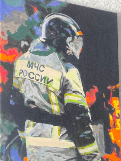 The image is a painting of a firefighter from the Ministry of Emergency Situations (MChS) of Russia. The firefighter is wearing a helmet and a reflective uniform with the "MChS ROSSII" text prominently displayed on a light yellow patch on their back. The background is abstract and depicts a chaotic scene with fiery orange and red elements, suggesting a fire or emergency situation. The painting style is impressionistic, with visible brushstrokes and blended colors. The location in the city of Psebai, Russia, is not explicitly depicted in the painting itself, but it is context provided with the image.