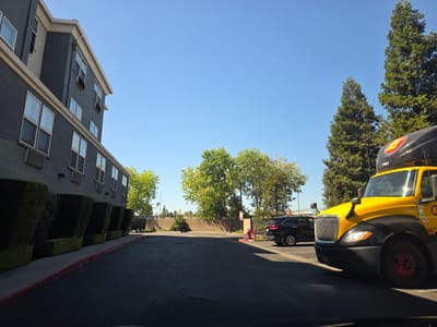 A yellow and black semi-truck is parked in a lot next to a grey building with a beige trim and numerous windows. The building appears to be a hotel or an apartment complex. A line of manicured, boxy hedges runs along the side of the building, separating it from a sidewalk. To the right of the building, a black SUV is parked. In the background, there are several tall, green trees and a brick wall. The sky is clear and blue, suggesting a sunny day. The time of day appears to be daytime, likely afternoon due to the shadows cast by the truck. The scene is an outdoor parking area, possibly at a commercial property. The truck has the number "79057" visible on its cab, along with a red and yellow circular logo. There is no activity or event taking place, and no people are visible in the image. The location is identified as West Sacramento, United States.