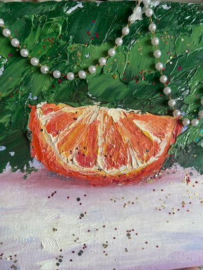 The image is a close-up of an artwork, possibly a painting or mixed media piece. The dominant colors are vibrant green, orange, and white, with scattered red, gold, and blue glitter. In the upper portion of the image, there is a thick, textured application of green paint, resembling foliage or branches. A string of pearl-like beads with a gold chain is draped across this green area.
The central subject is a thick, textured depiction of a slice of orange, rendered in shades of orange, yellow, and white paint. The texture is highly impasto, with visible brushstrokes and thick dabs of paint creating a dimensional effect. Red, gold, and blue glitter is scattered across the orange slice and in the surrounding green area.
The lower portion of the artwork features a textured white and light purple surface, also with scattered glitter. There is no indication of a specific city or location in Nachalovo, Russia, within the visual content of the image. The scene is static and appears to be a still life or abstract composition within the artwork itself. There are no people, actions, or interactions depicted.