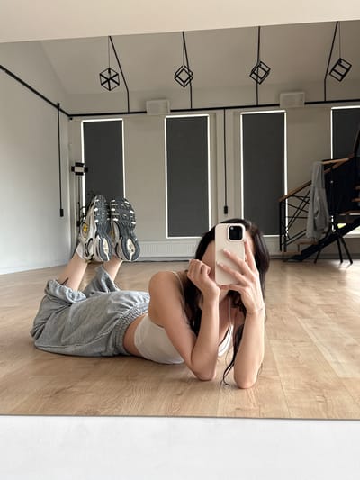 A full-body mirror selfie shows a young woman lying on her stomach on a polished wooden floor in what appears to be a dance studio or gym. She is wearing light grey sweatpants and a white sports bra, with athletic shoes on her feet. Her hair is dark and flows around her. She holds a light-colored smartphone in her right hand, obscuring her face as she takes the photo. In the background, the studio features large, dark-grey roller blinds covering windows, modern geometric pendant lights hanging from the ceiling, and a black staircase with railings. The overall environment is clean and modern. The location is identified as Agalarov Estate, Russia.