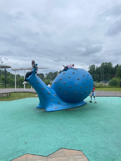 The image shows a playground scene in Sosnoviy Bor, Russia. The main subject is a large, blue, snail-shaped climbing structure with various grips. Several children are actively using the structure. One child in a tie-dye hoodie is climbing on the top part of the snail's shell. Another child, wearing a red shirt and white skirt, is standing on the ground near the snail's body. A third child, with long blonde hair and wearing a black top and dark pants, is on a lower part of the structure, seemingly descending or climbing.

The setting appears to be an outdoor recreational area with a green, soft-surface ground, typical of playgrounds. In the background, there are trees, a sports field with a net, and a cloudy sky. The weather seems overcast, suggesting it might be a cooler day, possibly late spring or summer. The overall mood is playful and active, with the children engaged in climbing and enjoying the playground equipment. There is no visible text in the image. The image is oriented sideways.