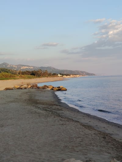 The image shows a serene beach scene in Marina di Casal Velino, Italy. The foreground is dominated by dark, textured sand, with a subtle ripple effect suggesting wind or receding water. Several large, rounded rocks are visible on the sand, acting as a natural barrier.
The midground features a strip of lighter-colored sand, leading to the gentle lapping of the sea. A lone individual is seated on the sand in this area, facing away from the camera.
Beyond the beach, a dense line of green vegetation, possibly pine trees and shrubs, forms a backdrop. Further inland, rolling hills rise, dotted with buildings that suggest a small town or village. The sky above is a pale blue, with scattered, soft clouds, indicating either early morning or late afternoon light. The sea is calm and expansive, with a subtle wave pattern near the shore, reflecting the soft hues of the sky. The overall impression is one of tranquility and natural beauty.