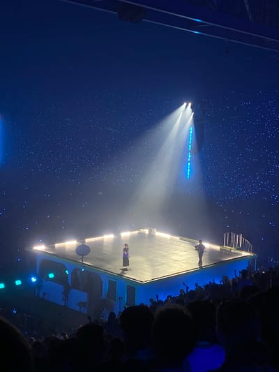 A concert is taking place in a dimly lit arena, with a stage that is lit by spotlights. The stage has a unique, futuristic design with a large, elevated platform that appears to be made of light-reflective material. Two performers are visible on the stage, one standing near the edge and the other further back. The audience is not clearly visible, but silhouettes of people can be seen in the foreground and on the sides of the image. The atmosphere is dark and atmospheric, with a generous scattering of small, sparkling lights that create a starry effect in the background, suggesting a magical or celestial theme. The stage is illuminated with bright white spotlights that create dramatic beams of light, and there are also blue lights visible on the stage and in the surrounding area, adding to the futuristic and ethereal ambiance. The overall impression is one of a grand, visually impressive performance with a focus on light and spectacle. The image appears to be taken from an elevated position, possibly from the stands, looking down at the stage and the crowd. The location is Arnhem, Netherlands, though no specific landmarks or text are visible to confirm this.
