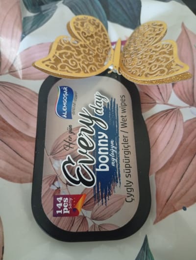 The image shows a close-up, top-down view of a package of wet wipes. The package is dark grey and rectangular with rounded corners, featuring a label with a floral pattern in shades of pink and green. The label prominently displays the words "Every day" in a large, stylized script. Above this, a logo for "ALEMGOŞAR" is visible. Below "Every day," the text "bonny" and "mylaym" are present. The side of the label reads "Çygly süpürgiçler / Wet wipes". In the bottom left corner of the label, there is a small square sticker with "144 pcs" and "Sany". Two decorative golden butterflies with intricate cut-out patterns are placed on top of the package, with one slightly overlapping the other. The background of the image is a light-colored fabric with a repeating pattern of large, stylized leaves in shades of pink and green. The overall impression is of a product display, possibly in a home environment given the fabric background, and not in Seýdi, Turkmenistan.