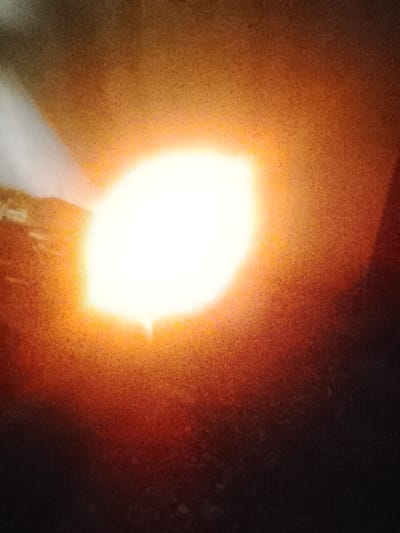 The image shows a bright, overexposed light source, likely the sun, against a dark, textured background. The colors are predominantly warm tones of orange, yellow, and white, with some darker reddish-brown and black areas. The overall impression is one of intense light and a possibly hazy or dusty atmosphere. There are no identifiable people, subjects, or specific objects visible that would allow for determining a location within the city of Orodo, Nigeria, or any other specific geographical context. The scene is abstract due to the overexposure and lack of clear details.