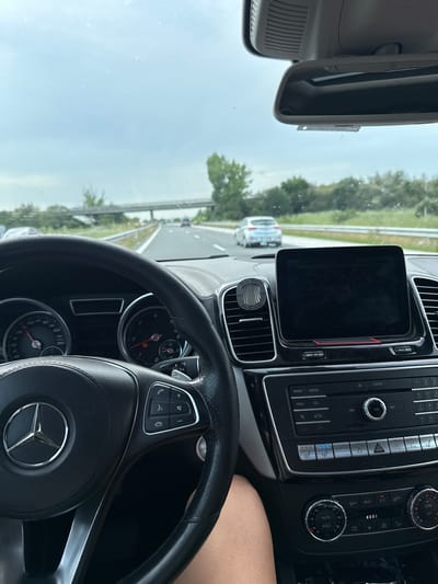 The image depicts the interior of a Mercedes-Benz vehicle, specifically the driver's perspective, on a highway in Gabarnitsa, Bulgaria. The steering wheel, adorned with the Mercedes-Benz logo, occupies the lower left quadrant, displaying multi-function controls on its right spoke. Behind the steering wheel, the instrument cluster shows a speedometer with the needle positioned between 100 and 120 km/h, and a tachometer with an illuminated warning light. To the right, a black infotainment screen is mounted on the dashboard, flanked by an air vent containing a cylindrical "Brooklyn Garage" air freshener. Below the screen, the center console features controls for radio, media, navigation, telephone, and climate control, with the heated driver's seat function visibly active (three blue lights). A driver's bare arm is partially visible in the bottom right corner.

Through the windshield, a multi-lane highway is visible under an overcast sky. The road is bordered by green trees and foliage. An overpass bridge crosses the highway in the mid-distance. Several other vehicles are present on the highway, with a silver hatchback discernible in the lane to the right, further ahead. A rear-view mirror is mounted at the top center of the windshield. The overall environment suggests driving on a rural or intercity highway during daytime.