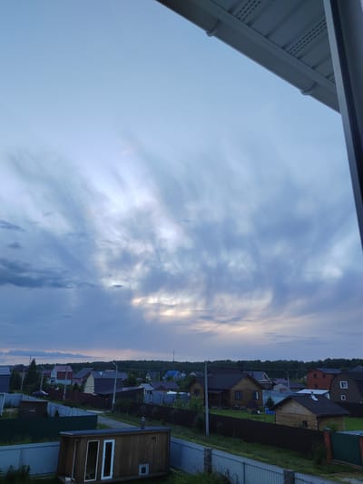 This media file depicts an outdoor scene of a village in Zhal'skoe, Russia, at dusk. The sky is dominated by dramatic, wispy clouds with streaks of light breaking through, suggesting either the last moments of sunset or the approach of twilight. The weather appears calm.

In the foreground, a simple wooden structure, possibly a small shed or sauna, with two white-framed doors is visible. It is situated within a fenced area, with more fencing and greenery extending into the middle ground.

Beyond the immediate foreground, a collection of residential houses with various roof colors and architectural styles are scattered across the landscape. Some houses are made of wood, while others appear to have more modern cladding. Fences of different materials, including metal and wood, delineate the properties. A dark line of trees forms the backdrop of the village, stretching across the horizon. A few streetlights are visible, hinting at the presence of roads and infrastructure. The overall mood is peaceful and rural, characteristic of a quiet evening in a village. There is no visible text in the image.