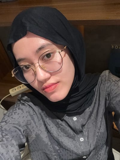 This image displays a single individual, identified as an adult female, situated indoors in Patrang, Indonesia. The subject is presented in a close-up, chest-up view, directly facing the camera with a neutral expression. She wears a black hijab, covering her hair and neck, and large gold-framed glasses with clear lenses. Her lips exhibit a glossy reddish tint. She is dressed in a dark gray long-sleeved collared shirt patterned with a subtle geometric design, featuring visible white buttons down the front placket. The background is dimly lit and features dark, indistinct surfaces, likely wood. A white multi-socket power strip with three visible outlets and an attached white cable is mounted on a lighter surface to the upper left. To the upper right, partially visible white capital letters "B O" are discernable on a dark surface, possibly text on a shelf or item. The scene is illuminated by a frontal light source.