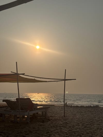 The image depicts a coastal scene in Bhagatwadi, India, during either sunset or sunrise. A bright, low-lying yellow sun, accompanied by prominent lens flare, dominates the upper-left quadrant of a hazy, light ochre sky. The sun's reflection creates a shimmering path of golden light across the calm ocean surface, which extends from the mid-ground to the horizon. In the dark sandy foreground, a rudimentary shelter is constructed from dark wooden poles supporting a multi-striped fabric canopy featuring red, orange, green, and yellow hues. Beneath this canopy, two dark wooden beach loungers are positioned, with a reddish fabric draped over one of them. To the far right, a small, dark, indistinct form rests on the sand near the waterline. At least one distant, dark vessel is discernible on the horizon line across the ocean. No explicit human actions or interactions are clearly depicted.