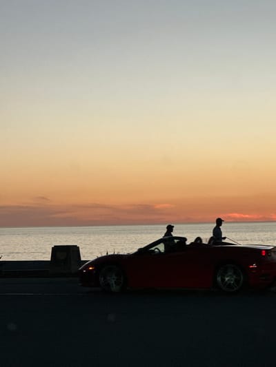 A convertible car is parked alongside a body of water, silhouetted against a sky transitioning from gray to a fiery orange sunset. Three figures are visible within the car, with one potentially standing. The car's wheels are detailed. The sky and water blend, with the horizon marked by a subtle color shift. The foreground consists of a dark surface, potentially a road or pavement.