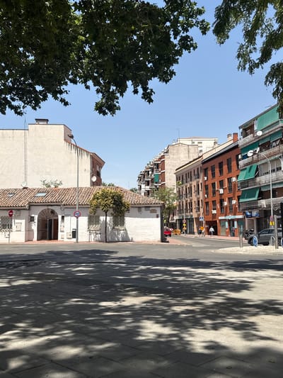 The image depicts a street scene in Alcalá de Henares, Spain, on a clear, sunny day. In the foreground, a paved area is covered with the dappled shadows of tree leaves, suggesting a tree-lined street or plaza.

To the left, a low, white building with a terracotta tiled roof and a prominent arched entrance is visible. The building has barred windows and several roof vents. Behind it, a tall, cream-colored building rises, its facade plain.

Dominating the right side of the image is a row of multi-story apartment buildings with brick facades. These buildings feature numerous balconies, many with green awnings extended. The ground floors appear to be commercial spaces. Air conditioning units are visible on the upper floors.

A street intersection is present, with cars and pedestrians in the mid-ground. A black car is parked near a traffic circle. Several traffic signs and streetlights are interspersed throughout the scene. Green trees are visible along the street and framing the top of the image. The sky is a clear, bright blue.