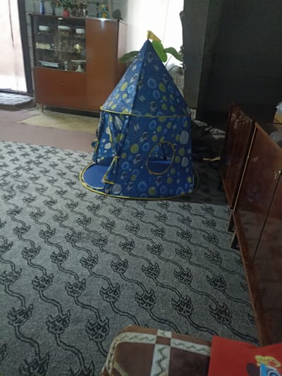 The image depicts an indoor scene, likely a living room, with a blue and yellow patterned play tent as the central focus. The tent has a playful design featuring astronauts, planets, and circular shapes. It is set up on a patterned grey carpet with a repeating floral or abstract motif. To the left of the tent, a wooden cabinet with glass doors displays various items, possibly decorative or collectible. To the right, a section of a wooden furniture piece, likely a cabinet or dresser, is visible. The background suggests a dimly lit environment, with part of a wall and a dark, possibly textured surface behind the tent. The scene is static and does not appear to capture any active events. There are no people visible in the image. The location is identified as Aygezard, Armenia.