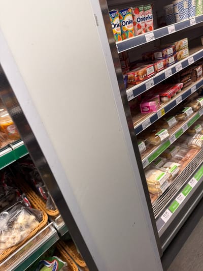 This is a photograph taken inside a retail environment, likely a supermarket, within Rotherham, United Kingdom. The image is taken from an elevated perspective, looking down into refrigerated shelves containing various food products.
The shelves are stocked with items such as "Onken" yogurt drinks, Tesco-branded products, "Potts & Bakers" desserts, and possibly other baked goods or food items packaged in clear plastic. Prices are visible on shelf labels, indicated in British pounds (£). The background suggests more chilled food sections, with baskets filled with items. The environment appears clean and well-lit. The overall setting is a typical retail grocery store.