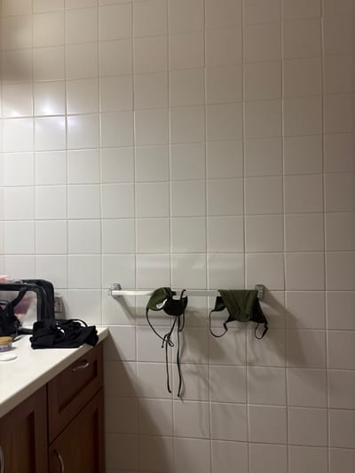 The image depicts an interior scene, likely a bathroom or utility area, within Olde Yard Village, British Virgin Islands. A white tiled wall, composed of square ceramic tiles with light-colored grout, occupies the majority of the frame. Attached horizontally to the wall, approximately one-third of the way up from the bottom, is a silver-toned metal towel bar. Two dark olive green fabric items, resembling swimwear tops with ties, are draped over this bar; one is on the left side of the bar, and the other is positioned to its right.
In the lower left quadrant, a section of a light-colored countertop is visible, extending from the wall. On this countertop, from left to right, are: a black pouch or bag with transparent sides, a crumpled piece of black fabric, a small circular jar with a light-colored lid, and a white circular cotton pad. Beneath the countertop, a portion of a brown wooden cabinet or vanity unit is visible, featuring a drawer and a cabinet door, both equipped with silver-colored pull handles. The scene is illuminated by an unseen light source, casting soft shadows beneath the hanging garments and reflecting off the tiles. No individuals or active interactions are present.