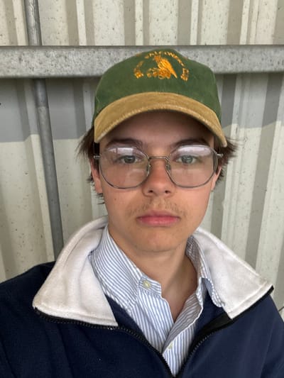 A young man, wearing a dark blue fleece jacket over a light blue and white striped collared shirt, is looking directly at the camera. He is wearing a green and brown baseball cap with gold embroidery. He has dark brown, slightly messy, shoulder-length hair and is wearing glasses with a silver frame. He appears to be in a small, possibly enclosed area, as evidenced by the corrugated metal wall in the background, possibly located near a farm in the city of South Ribble, United Kingdom, based on the provided coordinates.