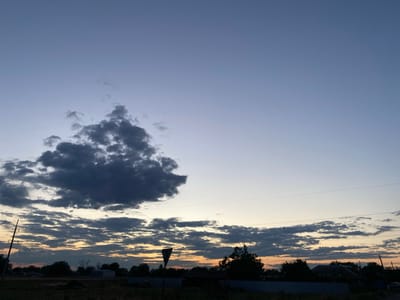 This is a photograph of a rural landscape at dusk in Taganrogsky, Russia. The sky transitions from a pale blue at the top to a soft orange hue near the horizon, indicating the setting sun. A large, dark cloud formation dominates the upper left portion of the sky, while a band of smaller, textured clouds stretches across the mid-ground.
The foreground is dark and appears to be a field of dry grass and low vegetation. Silhouetted against the horizon are trees, some residential structures, and a power line with a triangular warning sign on a pole. The overall mood is tranquil and serene, characteristic of the end of a day. The location is suggested by the power lines and rural architecture typical of the region. No visible text is present in the image.