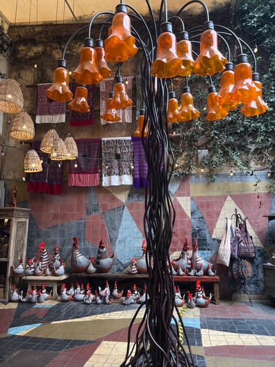 The image showcases an eclectic indoor market or courtyard. Woven textiles hang vertically, interspersed with decorative lamps. On the right, shelves display whimsical ceramic bird figurines in orderly rows. Above, a striking chandelier with orange, flower-like shades contrasts with strands of industrial cables. The floor exhibits a geometric tile pattern with triangles of varying colors, adding to the space's unique aesthetic. Vegetation climbs up a wall on the left.