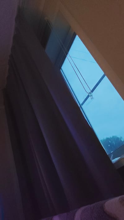 The image displays a window with a blue tint, likely from an overlay or the sky. A beaded chain hangs from the top of the window frame, attached to a small white mechanism. To the left of the window, dark purple curtains are drawn. In the foreground, a blurry surface with purple and cream stripes is visible, possibly a blanket or upholstery. The overall scene is dimly lit, with the bright window providing the primary light source. The image is taken from a low angle, looking upwards towards the window. There is no indication of specific location within Redkino, Russia, beyond the fact that the image is presented in that context.