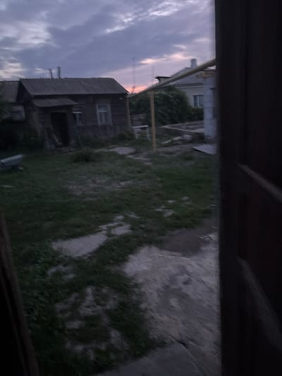 The image depicts a rustic backyard scene at dusk in Novoanninskiy, Russia. A weathered wooden house with a corrugated metal roof stands prominently in the left portion of the frame. A small porch with steps leads to a dark doorway. To the right of the wooden house, a lighter-colored, possibly brick or concrete, building is partially visible, with a green bush in front of it. The ground is covered with patchy grass and concrete slabs, suggesting a utilitarian rather than decorative outdoor space. In the distance, a faint pink and purple hue in the cloudy sky indicates the setting sun. A single metal structure, possibly a utility pole or antenna, is visible against the sky. A wheelbarrow can be seen on the grass to the left of the wooden house. The foreground is dominated by a dark, out-of-focus doorway, framing the view of the backyard. The overall impression is one of a rural or semi-rural dwelling, with signs of age and modest upkeep.