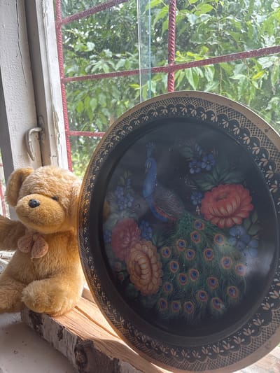 A teddy bear sits on a piece of birch wood next to a decorative black tray with a painted peacock and flowers. The scene is set on a windowsill with a view of green foliage through a red-barred window. Raindrops are visible on the windowpane. The location is identified as Golenishchevo, Russia.