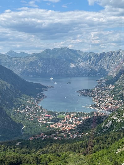 The image presents an aerial view of Kotor Bay in Montenegro, specifically capturing the area around the city of Mirac. The landscape is dominated by the bay's blue waters, surrounded by imposing mountains. A settlement, likely part of Mirac, is visible along the coastline, with clusters of buildings interspersed among the greenery. The scene is bathed in natural light, with skies featuring scattered clouds. Several boats dot the bay's surface.