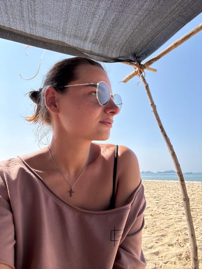 This image captures a serene scene of a woman relaxing on a sunny beach, viewed from a low-angle perspective. The woman, who appears to be lying down, is looking upwards with a calm expression. She has dark hair styled in a bun and wears round, gold-rimmed sunglasses. Her attire includes a light brown/mauve off-the-shoulder top, with a black bikini strap visible underneath. A delicate silver chain necklace with a small cross pendant rests on her chest.
She is partially shaded by a simple, makeshift structure composed of dark, mesh-like fabric supported by rough wooden sticks or branches. The background reveals a sandy beach extending towards the ocean, which appears calm under a clear blue sky. Bright sunlight illuminates the scene, suggesting it is daytime with good weather.
A small, stylized graphic resembling an incomplete rectangle is subtly visible on the right sleeve of her top. The overall atmosphere conveys relaxation and enjoyment of the beach environment. The provided location context for this media file is Bhagatwadi, India.