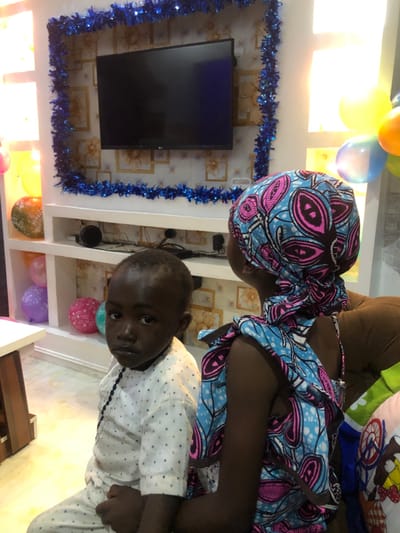 The scene depicts an interior room, located in Barakin Acha, Nigeria, decorated with festive elements. A young, dark-skinned child, identified as male, is positioned in the left foreground, wearing a white long-sleeved shirt with a subtle dot pattern and black beaded ornamentation on the front. He looks directly towards the camera. To his right, with her back to the camera, is a second individual, wearing a patterned headwrap and a sleeveless dress in blue, pink, and brown African print. This individual is seated on brown upholstered furniture.
The background features a wall with a subtle patterned wallpaper. A wall-mounted flat-screen television, branded "LG," is centrally placed, framed by blue tinsel garland. Below the television, a white shelving unit contains speakers and other electronic components. Multiple inflated balloons in yellow, orange, blue, pink, and purple are visible throughout the background. A partial view of a light-colored table is present in the lower-left corner.