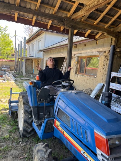 A person is sitting on a blue Landini tractor in what appears to be a rural or semi-rural environment. The tractor is parked on grassy land, near a building under construction. The building is partially exposed and appears to be a two-story structure, with the upper portion freshly plastered or painted. Construction materials are visible near the tractor. The image has elements of an agricultural setting combined with renovation activity. The location is Rish, Bulgaria.