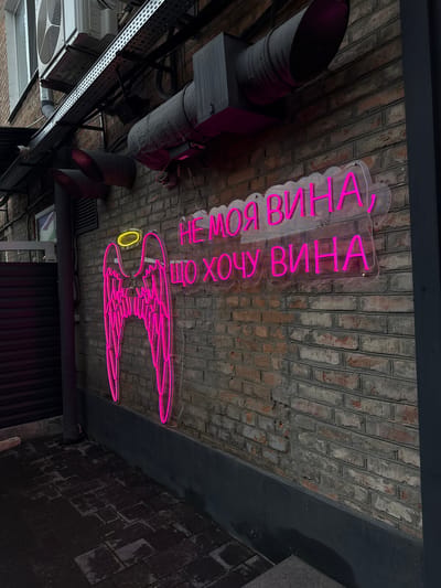 An outdoor brick wall in Zviahel, Ukraine, is depicted under dim ambient light, illuminated by two neon signs. The wall, constructed of weathered red and grey bricks, features various utility installations. In the upper left quadrant, a white air conditioning unit is mounted, with a network of metal cable trays and pipes extending horizontally and vertically across the wall. Below the air conditioner, two dark grey, funnel-shaped ventilation ducts project outwards. A small, white security camera is visible on the left side of the wall.
Two prominent neon signs are centrally positioned. On the left, a large neon sign in bright pink outlines a pair of angel wings, topped with a yellow neon halo. To its right, another pink neon sign displays Cyrillic text on a clear acrylic backing, shaped like a cloud or speech bubble. The text reads: "НЕ МОЯ ВИНА, ЩО ХОЧУ ВИНА" (transliterated as "NE MOYA VINA, SCHO KHOCHU VINA"), which translates to "It's not my fault that I want wine," utilizing a pun on the word "вина" (meaning both "fault" and "wine").
The ground beneath the wall consists of dark-colored, rectangular pavers, appearing damp or wet. A dark vertical structure, possibly a pillar or building edge, is visible on the far right. Ventilation ducts, one large and curved on the upper right, and associated piping are also affixed to the brickwork. The overall scene suggests an urban commercial or entertainment establishment.