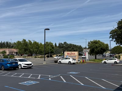 The image captures a sunlit commercial parking lot on a clear day in Antelope, United States. The sky is a light blue with wispy, streaky white clouds, indicating midday. No people are discernible in the scene.

In the foreground, a dark asphalt parking lot features blue and white painted parking lines, including a handicap parking symbol. On the left, a blue Kia Forte is parked behind a white Honda Civic. The Honda Civic's license plate appears to be "9LJZ285", and the Kia Forte's "9K95325". Several black street lights are positioned throughout the lot.

Towards the mid-ground, a prominent sign displays "BEL AIR" in a large, red, stylized font, with "24 HOUR FITNESS" written beneath it in black. The numbers "6887-7909" are vertically printed on the left side of the sign. A white Toyota Prius is parked near this sign, and a red fire hydrant with a yellow pole is visible beside it.

Beyond the parking lot, a tree-lined road features a white Subaru Impreza hatchback driving, with other vehicles visible in the distance. Green trees and foliage are abundant, bordering the parking lot and road, and low-rise commercial buildings can be seen in the background. A small red 'STOP' sign is also visible in the distance. The overall setting is a typical, quiet commercial shopping center during normal business hours.