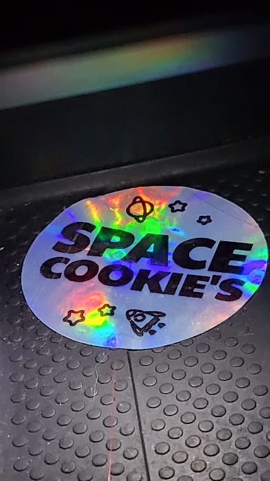 A circular holographic sticker is positioned on a dark, textured surface. The sticker features the black uppercase text "SPACE COOKIE'S" and outlines of celestial objects: a bell-shaped planetoid, multiple stars, and a rocket ship. Iridescent rainbow light reflects across the sticker's surface. The underlying surface is dark grey or black, characterized by a repeating pattern of raised circular nubs, consistent with an anti-slip mat or flooring. A subtle linear indentation or mark runs vertically through the lower half of the sticker. Fine, light-colored fibrous debris is visible on the textured surface adjacent to the sticker. The upper portion of the image is dark, exhibiting a distinct horizontal band of diffused rainbow light. This scene is located in Magaluf, Spain.