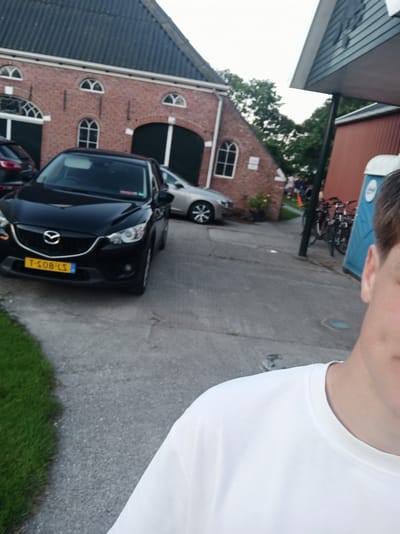 The image, taken at the Westereems 10, 9981 AD Uithuizen, Netherlands, captures an outdoor scene with a focus on a black car and part of a person. The car, a Mazda CX-5, is parked on a concrete surface, its yellow license plate clearly visible with the registration T-S08-LS. Behind the car, a brick building with arched windows and dark green doors suggests a rural or industrial setting. To the right of the car, a blue portable toilet and several parked bicycles are visible, indicating a possible event or gathering. The background shows greenery and trees, with a glimpse of other people in the distance. The foreground features the shoulder and neck of a person wearing a white t-shirt, suggesting the photo was taken from a first-person perspective. The sky is overcast, suggesting an early evening or late afternoon time.