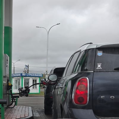 The image depicts a black Mini Cooper parked at a gas station in what appears to be Дорскі сельскі Савет, Belarus, based on the Cyrillic script on a nearby sign. The car is positioned near a gas pump. A motorcycle is parked to the side of the gas pump. A building with "СТРАХОВАНИЕ" (Insurance) written on the side in Russian is behind the pump. The sky is overcast.