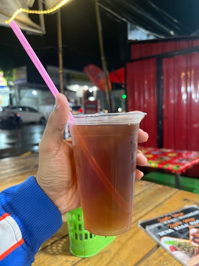 A person's hand, wearing a blue jacket with white and red trim, holds a plastic cup filled with iced tea and a pink straw. The cup is condensation-covered, suggesting it is cold. The hand and cup are positioned in the foreground, on the right side of the frame, obscuring part of a wooden table. In the background, there is a blurred red building with corrugated metal siding and what appears to be an outdoor seating area with a colorful tablecloth on a table. The scene is dimly lit, with strings of yellow lights overhead and streetlights visible in the distance, indicating it is nighttime. Cars and buildings are also visible in the blurred background, suggesting an urban or semi-urban setting. A green plastic container is partially visible on the table near the cup. A menu is also visible on the table in the lower right corner, with text that appears to be in Indonesian. The overall setting suggests a casual dining or street food establishment in Boja, Indonesia, possibly an outdoor food stall or restaurant.
