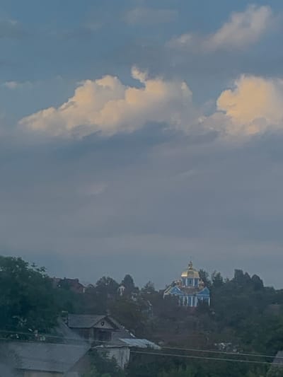 The image displays a daytime view of the Church of the Dormition of the Blessed Virgin Mary in Deliatyn, Ukraine. The church is positioned prominently on a tree-covered hill in the midground, featuring a central golden dome and light blue walls with white architectural details. Dense green foliage surrounds the church, with other residential buildings partially visible among the trees on the hillside. In the foreground, the upper sections and dark roofs of additional structures are discernible, partially obscured by more dark green foliage. Horizontal lines, consistent with utility wires, traverse the lower portion of the frame. The sky occupies the upper half of the image, presenting a mix of light blue and various cloud formations, including bright, sunlit cumulus clouds in the upper sections and more muted, greyish clouds filling the expanse below them. No individuals or active interactions are visible within the scene.