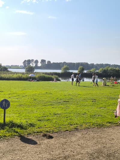 The image depicts a sunny day at a body of water, likely a beach or lakefront, in Broek op Langedijk, Netherlands. In the foreground, a grassy area slopes gently towards the water. A blue and white striped umbrella shades a group of people lounging on the grass. Further down, near the water's edge, several individuals are standing and interacting, with one person possibly playing with a soccer ball. A yellow trash receptacle stands on the grass to the right, and a picnic table with people seated is visible beyond it. The background shows a line of trees and vegetation along the far bank of the water, with a clear blue sky overhead. A sign with a pedestrian icon is present on the left side of the image, indicating a walking path. The overall atmosphere is one of a relaxed recreational setting.