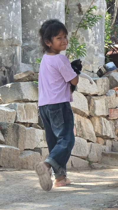 In Ba Ngoi Ward, Vietnam, a young child with black hair stands on stone steps, holding a black and white cat. The child wears a light purple shirt and blue jeans, with tan sandals on their feet. The background includes stone structures and greenery.