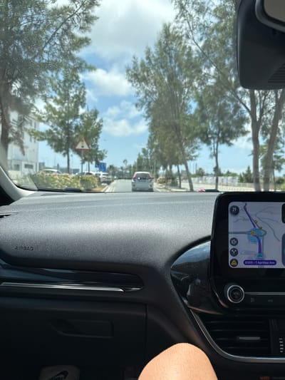 The image shows a view from the inside of a car, looking forward through the windshield. The car is on a paved road lined with trees, and there are buildings and other cars visible in the distance. The dashboard of the car is in the foreground, with a navigation screen displaying a map and route. The navigation screen indicates the location as "E305 - 1 Apriliou Ave", which is a street in Paralimni, Cyprus. The sky is partly cloudy, and the overall lighting suggests it is daytime. A person's leg is visible in the lower right corner of the image.