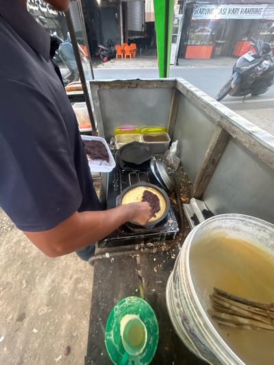 A vendor in Blanggunci, Indonesia, prepares a dish on a portable stove. The vendor, seen from the shoulders down, wears a dark blue shirt and is in the process of spreading a dark red filling onto a thin, golden-brown crepe cooking in a black pan. The crepe is on a two-burner gas stove that appears to be part of a food cart. To the left of the stove, a white rectangular container holds more of the dark red filling. Above the stove, two yellow containers with lids are visible, and behind them, a plastic bag contains more ingredients. To the right of the stove, a large white bucket is filled with a light yellow liquid and several wooden sticks. The food cart itself is constructed of gray metal sheeting and wooden beams. In the background, a street is visible with parked motorcycles, orange chairs stacked outside a building with a sign reading "WARUNG NASI KARI KAMBING". The ground around the food cart is paved with a rough, gray material. The scene suggests an outdoor food stall operating on a street.