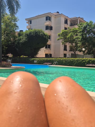 A first-person perspective shot shows a person's legs, glistening with water droplets, positioned in the foreground. The legs are tanned and appear to be resting on a surface that is slightly out of focus. Behind the legs, a bright blue swimming pool is visible, its clear water reflecting the sunlight and creating a vibrant turquoise hue. The pool area is bordered by a light-colored tiled edge. In the mid-ground, a manicured green hedge runs along the perimeter of the pool area, punctuated by wooden benches. Further back, a large, multi-story building with a light-colored facade and dark-shuttered windows stands under a clear blue sky. Lush green trees with dense foliage and some with purple flowers frame the building. A tall palm tree's fronds are visible on the far left side of the frame. The overall scene suggests a sunny day at an outdoor swimming pool, likely in a residential or hotel complex in Les Meravelles, Spain.