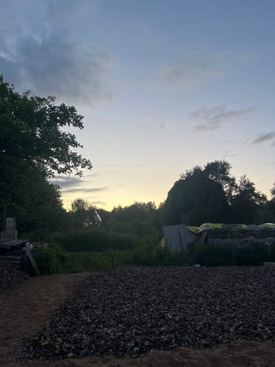 The image depicts an outdoor scene at dusk, possibly in the village of Klyobushnevo, Russia. In the foreground, a large pile of dark grey gravel is visible, with a section of lighter sand next to it. A wooden stake is partially embedded in the gravel. Behind the gravel, there is a dense area of green foliage and trees. In the mid-ground, a dilapidated structure with a yellow tarp is visible, and further back, a small, illuminated house with a steeply pitched roof can be seen amongst the trees. The sky above is a gradient of pale blue to light yellow, with scattered dark and light clouds. Power lines are stretched across the upper part of the image. The overall impression is one of rural or semi-developed land at the end of the day.