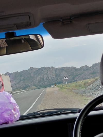 The image shows a view from inside a car driving on a highway in Қапанбұлақ ауылдық округі, Kazakhstan. A large, rocky mountain range dominates the background. The highway stretches ahead, with a white line marking the lanes. To the right of the road, a triangular road sign with a black arrow pointing to the right is visible. In the foreground, the steering wheel of the car and a dashboard are visible. A crumpled purple plastic bag is placed on the dashboard. A large truck is partially visible on the left side of the road. The sky is overcast.