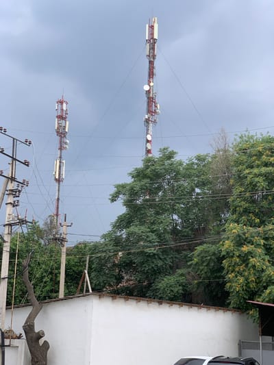 The image depicts two tall telecommunication towers, one on the left and a taller one on the right, against a cloudy sky. Both towers are lattice structures, painted red and white, equipped with multiple antennas and dishes. Below the towers, a white wall is visible, topped with a corrugated roof. Green trees with lush foliage surround the lower parts of the towers and the wall. Power lines and utility poles are also present, crisscrossing the scene and connecting to the towers. In the foreground, a gnarled tree trunk with cut branches extends upwards. A small bird is perched on top of the white wall. The scene is set in the city of Urtaaul, Uzbekistan.