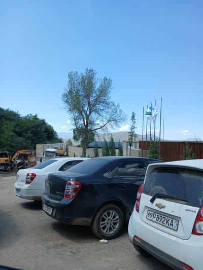 The image captures a sunny day in an outdoor setting, possibly in Uzbekistan, given the license plates. Two cars, a white Chevrolet Spark with license plate "95 P892KA UZ" and a dark-colored car behind it with license plate "367 284 BB", are parked side-by-side. The white car's rear is facing the viewer, revealing its license plate and "SPARK" badging. Behind the cars, a building with a wooden facade and glass windows is visible, along with several flagpoles flying flags. The scene also includes trees, hills, and a clear blue sky. Further in the background, construction equipment, including an excavator and a truck, are present, suggesting ongoing work or development. The overall atmosphere appears calm and ordinary, with no specific activity or event prominently featured. The lighting suggests it's daytime.