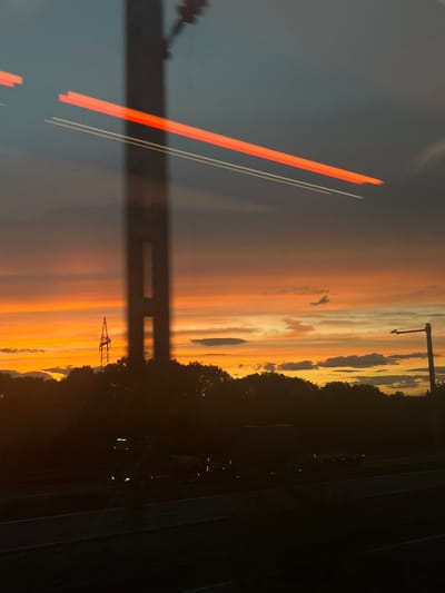 The image depicts a sunset scene with a dramatic sky painted in hues of orange, yellow, and gray. In the foreground, a dark silhouette of trees borders a highway where several vehicles, including a truck, are visible. A tall, dark pole-like structure, possibly a utility pole or a part of a bridge, bisects the image vertically, with blurry red and white streaks of light, likely from passing vehicles or reflections, overlaying it. In the distance, a lattice-like tower, characteristic of power lines, stands against the vibrant sky. The location is identified as Bubenreuth, Germany.